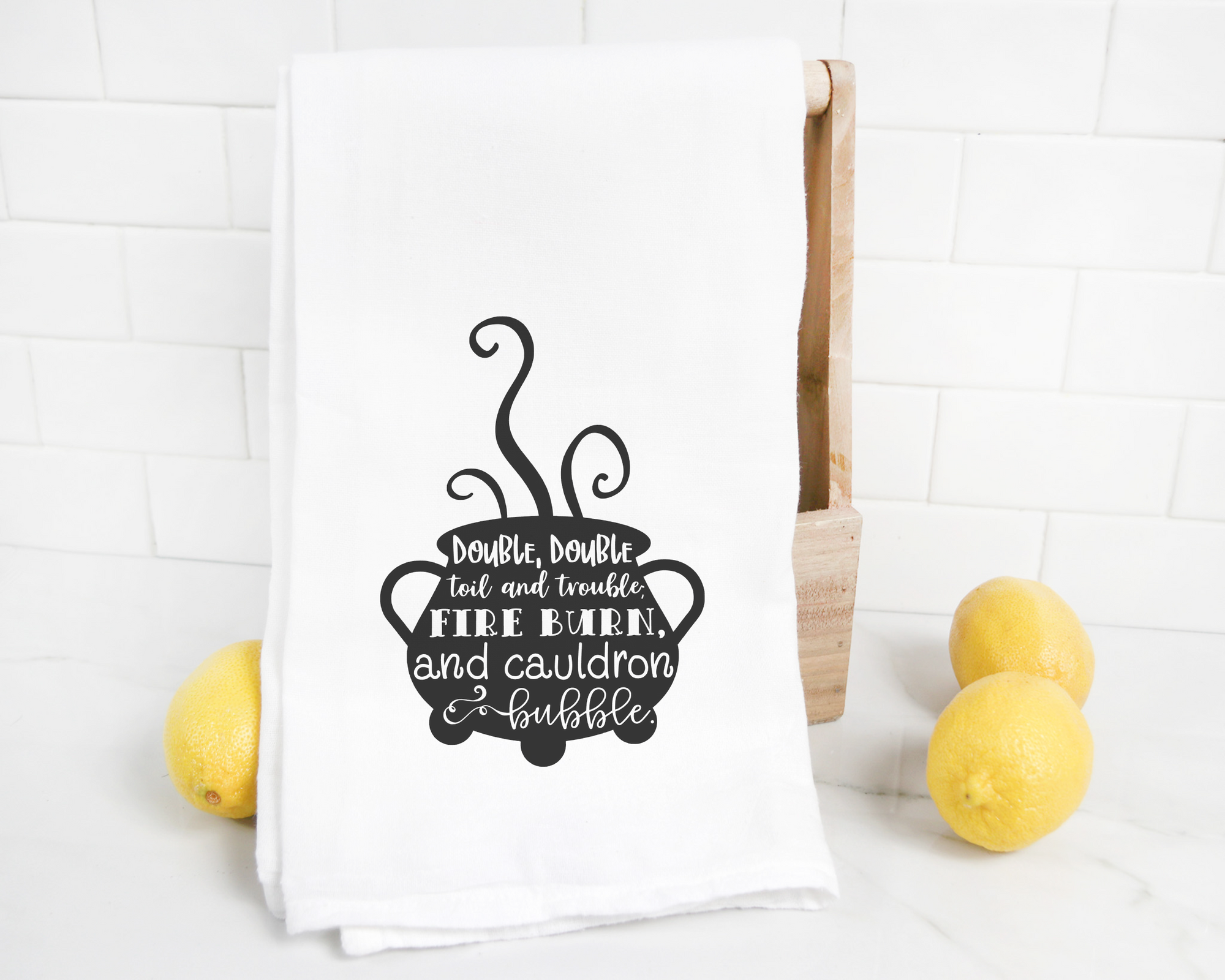 A white Double Double Toil and Trouble Tea Towel, displayed on a wooden stand with two lemons beside it, perfect for Halloween