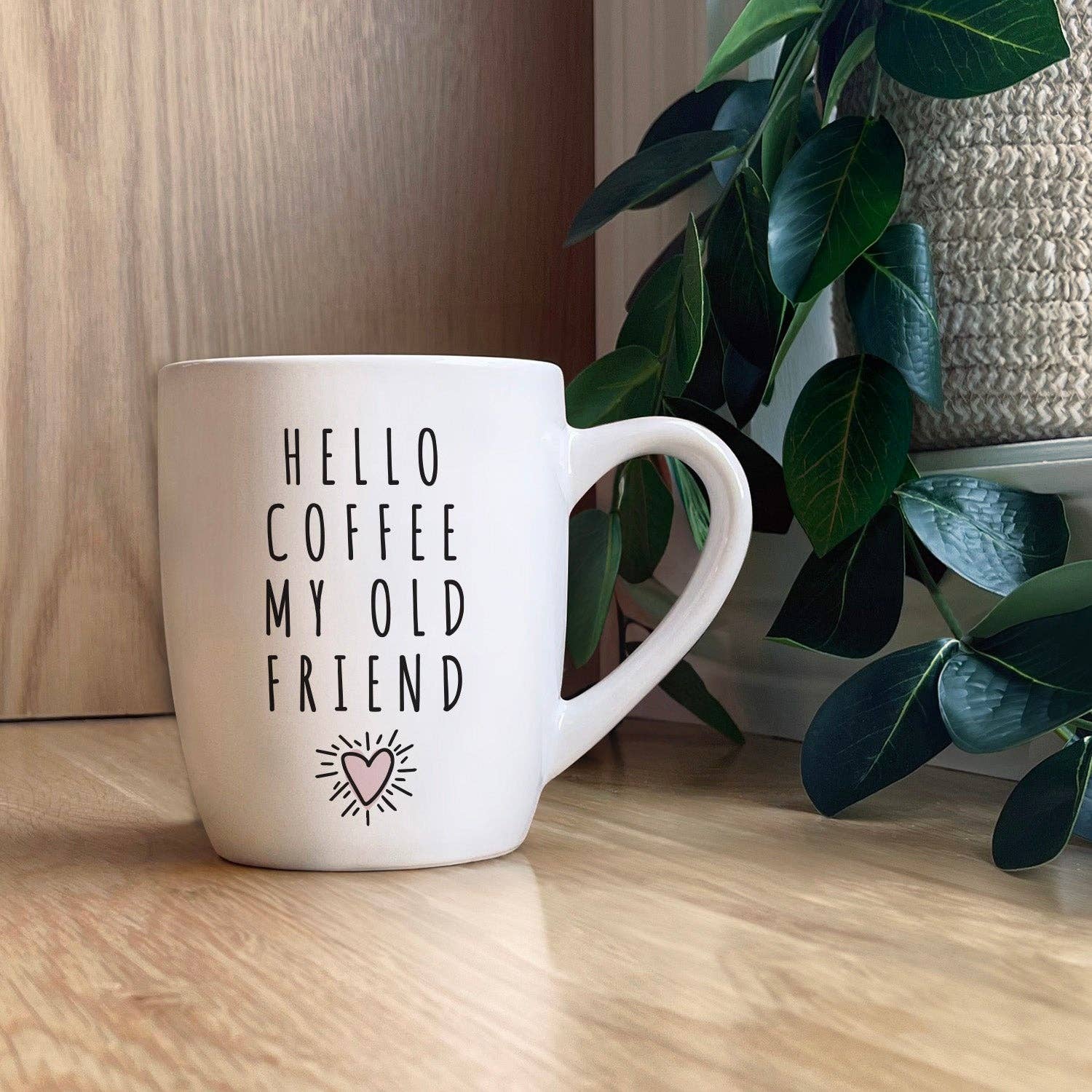 The "Hello coffee, my old Friend" white coffee mug, adorned with the text “HELLO COFFEE MY OLD FRIEND” and featuring a small heart and sunburst drawing, sits on a wooden floor beside a green plant and some cozy towels.