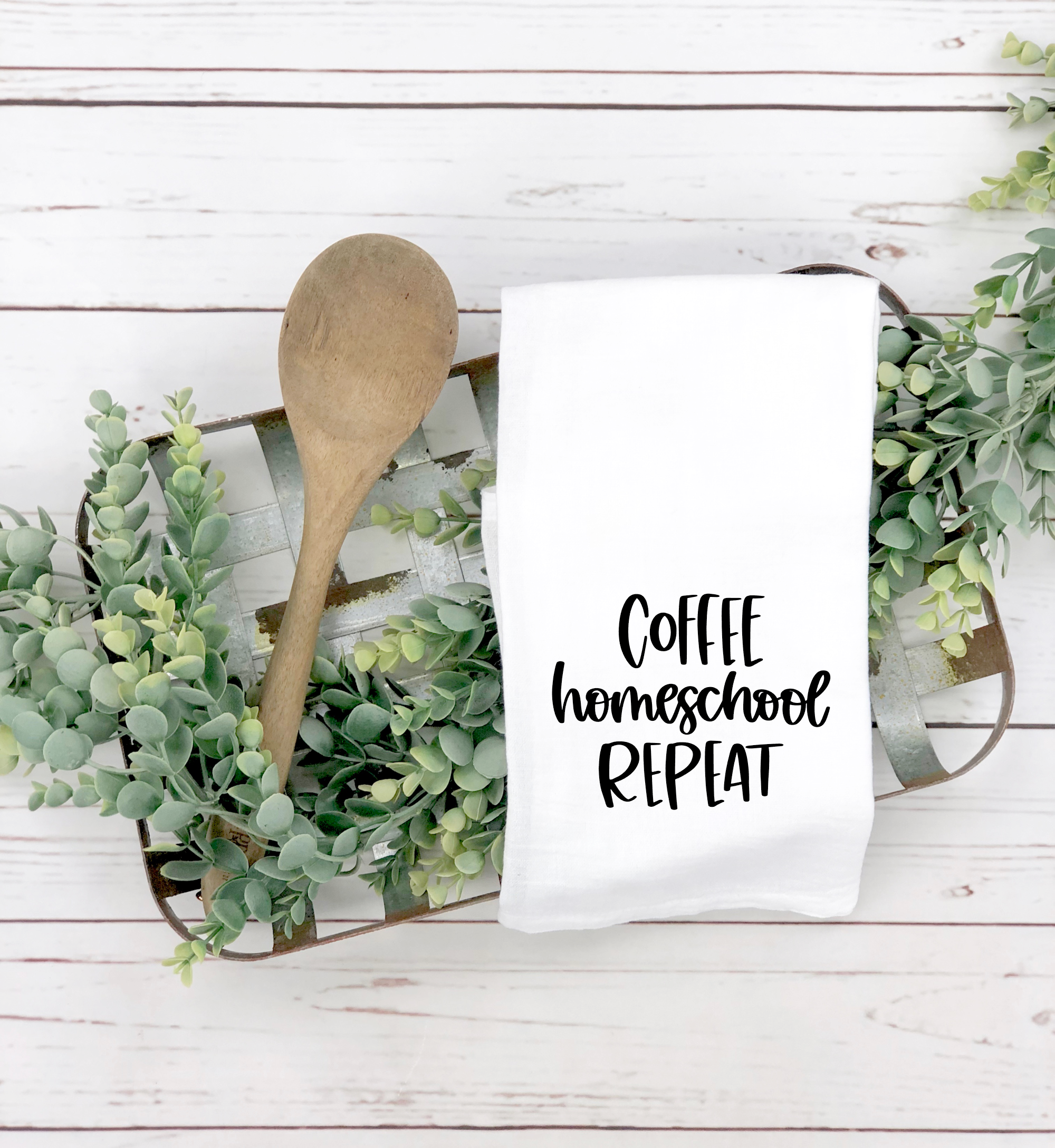 A perfect back-to-school gift is the Coffee Homeschool Repeat Tea Towel, paired with a wooden tray, wooden spoon, and greenery. The white kitchen towel features the phrase "Coffee Homeschool Repeat" in black text.