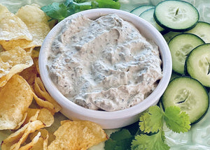 A bowl of creamy jalapeno ranch dip surrounded by potato chips, cucumber slices, and cilantro leaves.