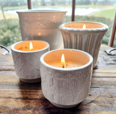 Four Eclectic Small Fluted Pot Swan Creek Candles in white ceramic are lit and arranged on a wooden surface, with an outdoor view through the window in the background.