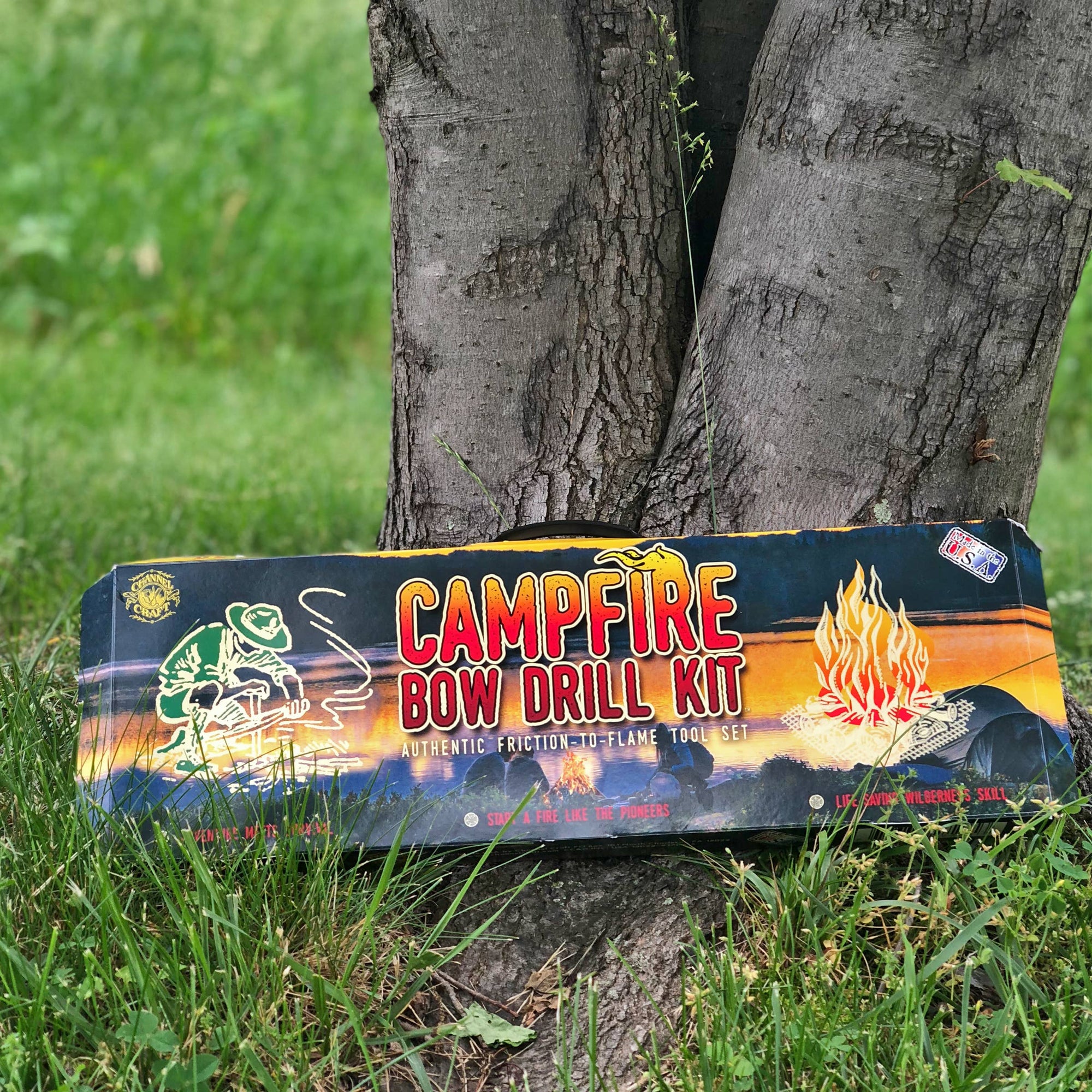 Campfire Bow Drill Kit placed at the base of a tree, packaging features vibrant fire graphics and text.