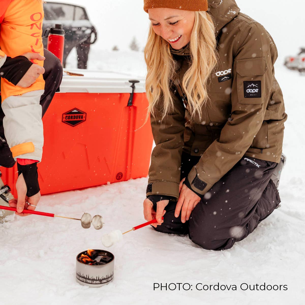 A woman in winter attire roasting marshmallows over a City Bonfires® by Coleman® in the snow, with an orange cooler and another person partially visible.