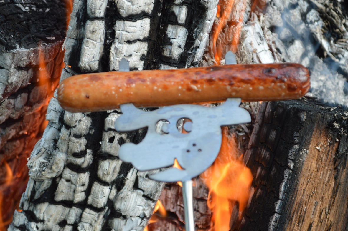 A sausage cooking over an open fire, held by an Animal Roaster with a decorative handle shaped like a smiling cartoon face.