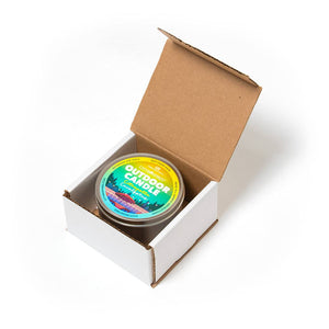 A small open cardboard box containing a colorful tin of The Outdoor Candle - Citronella and Eucalyptus on a white background.