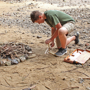 A boy in a green t-shirt and khaki shorts kneels on sandy ground, using a Campfire Bow Drill Kit to start a fire near a pile of small rocks and twigs.