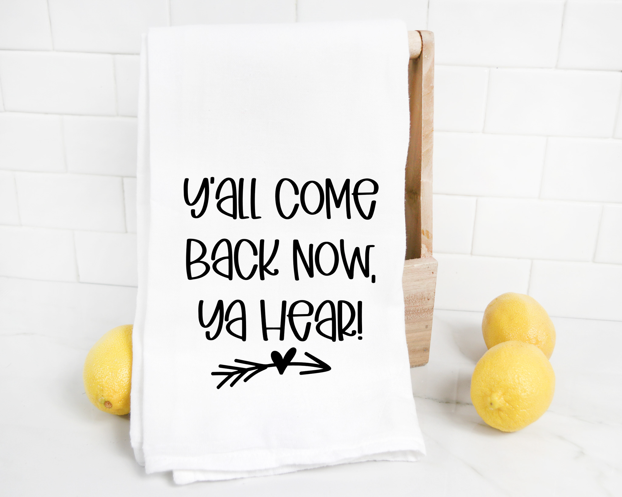 The "Y'all Come Back Now Ya Hear Tea Towel," featuring the charming phrase in black font, is displayed on a wooden stand with three lemons nearby, making it an ideal housewarming gift.