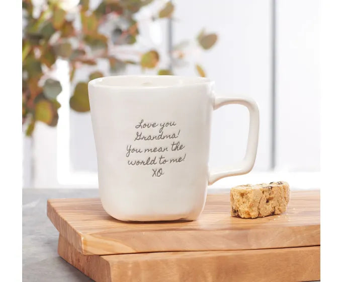 A Studio M coffee mug from the Heart Notes collection, featuring a cookie and resting on a wooden cutting board.