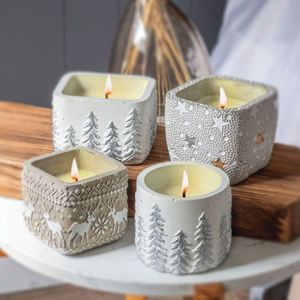 Four Winter Wonderland Small Round Pot Swan Creek Candles in textured gray holders, featuring winter-themed designs like trees, stars, and reindeer, are arranged on a wooden surface.