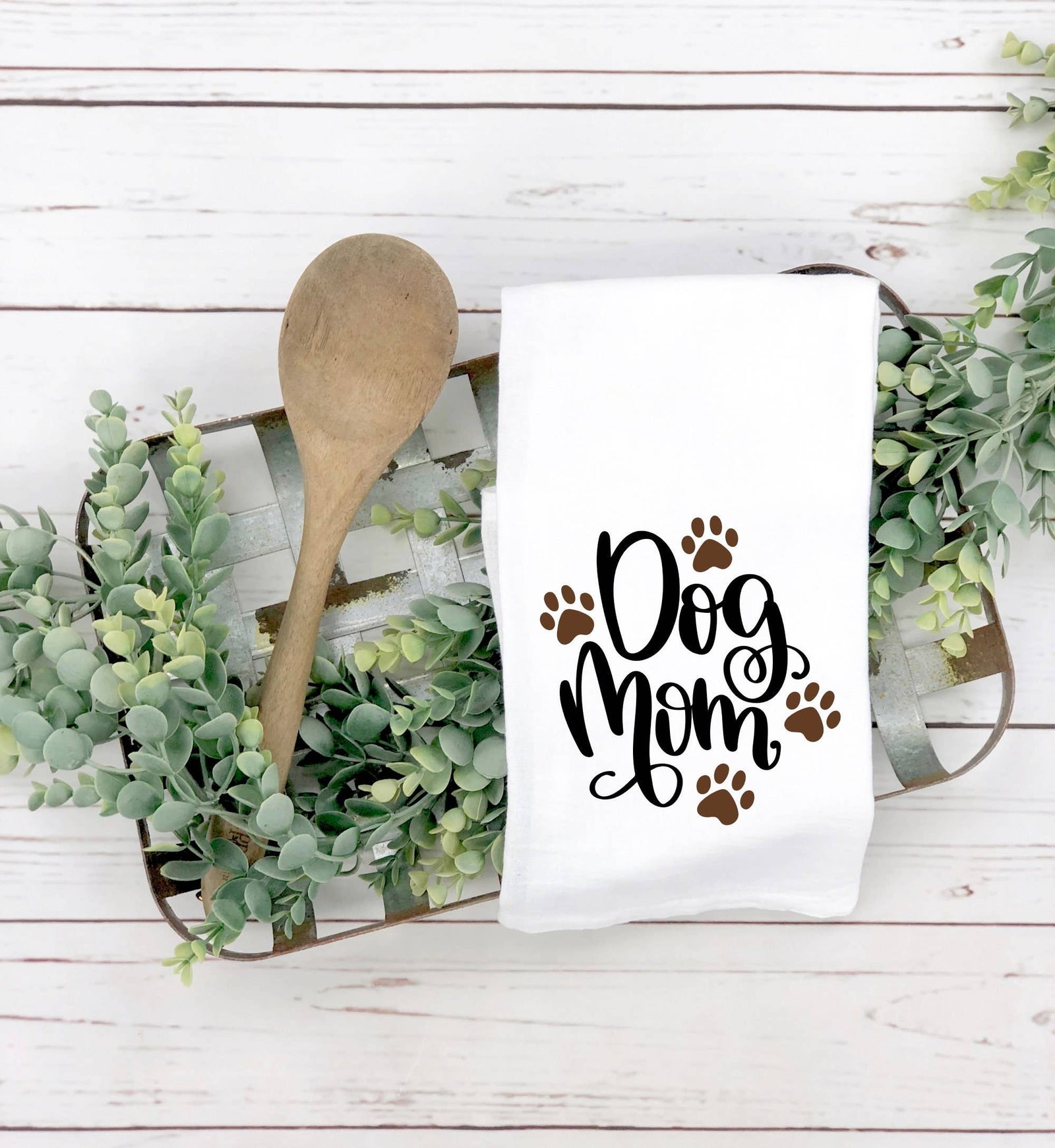 A wooden tray with a wooden spoon, faux greenery, and a discontinued "Dog Mom" tea towel with paw prints—an ideal, unique gift for dog lovers.
