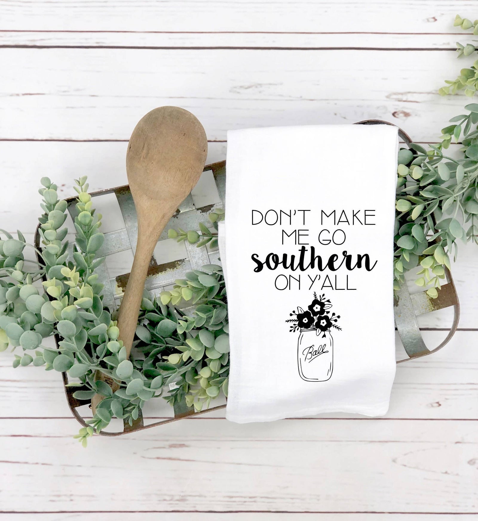 A wooden spoon and the discontinued "Don't Make Me Go Southern on Y'all" tea towel are displayed on a tray with green foliage—a charming kitchen gift idea.