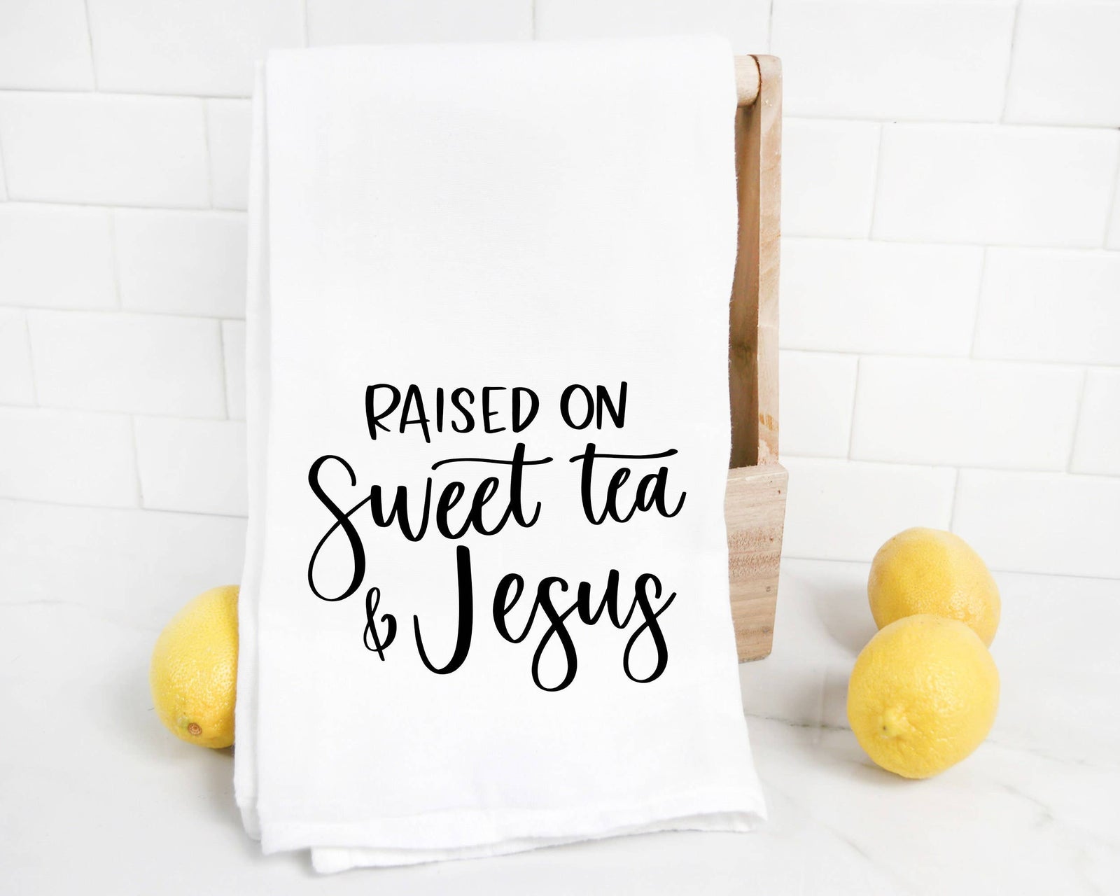The discontinued "Sweet Tea and Jesus Tea Towel" features “Raised on sweet tea & Jesus” in black script, made with heat transfer vinyl, displayed over a wooden holder next to three lemons on a white surface.