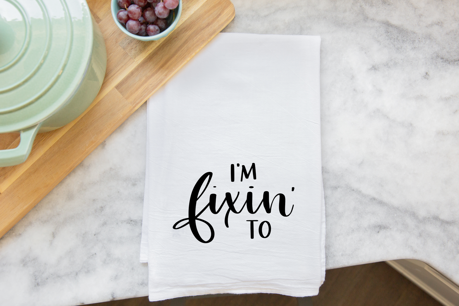 The discontinued "I'm Fixin' To Tea Towel" features the phrase in black cursive and block letters on white fabric, displayed on a marble countertop beside a green pot and grapes—a humorous Southern gift for any home.
