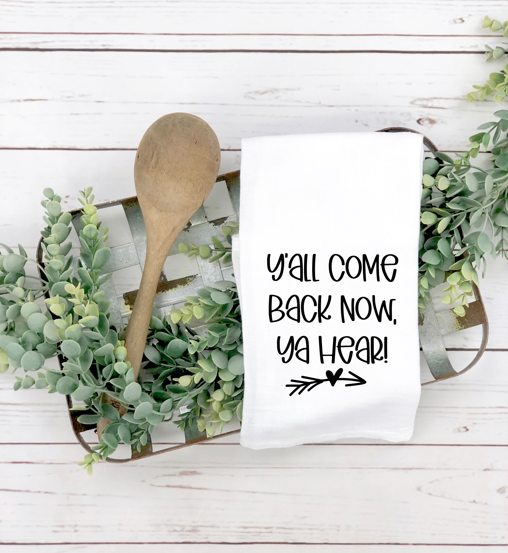 A wooden spoon and the "Y'all Come Back Now Ya Hear Tea Towel" with the charming text "Y'all come back now, ya hear!" are arranged on a tray adorned with green foliage on a white wooden surface, creating an ideal housewarming gift.