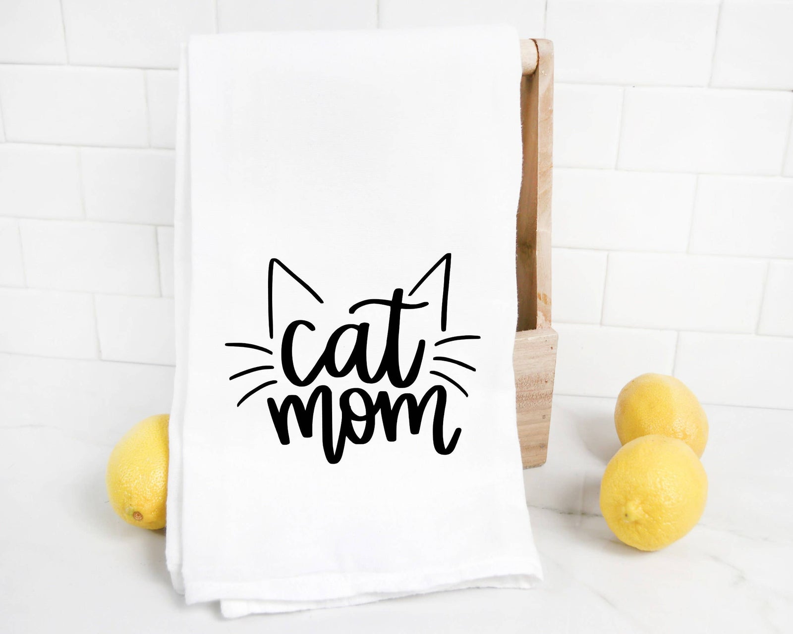 The Cat Mom Tea Towel DISCONTINUED features "cat mom" and cat ear graphics in black on white, shown on a wooden holder by three lemons—an ideal cat lover's kitchen gift.