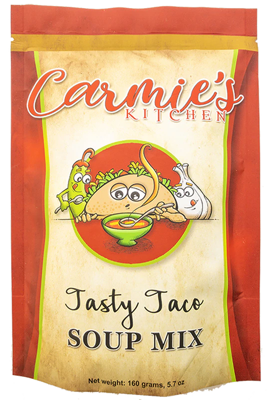 A package of Tasty Taco Soup Mix from Carmie's Kitchen features a whimsical illustration of a taco, avocado, and onion on the front, accompanied by the text "Tasty Taco Soup Mix" and indicating a net weight of 160 grams (5.7 oz). This mix is ideally combined with ground beef and a variety of spices to craft a deliciously savory taco soup.