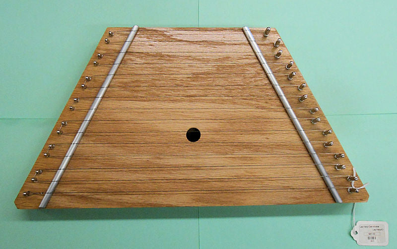 An acoustic folk instrument, specifically a triangle-shaped wooden instrument by TK O'Brien, hanging on a wall. The Lap Harp - Oak Top.
