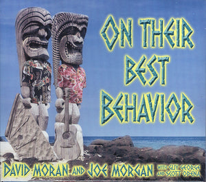 Two tiki statues in Hawaiian shirts are positioned on a rocky shore with the ocean as the backdrop. The text reads "On Their Best Behavior by David Moran and Joe Morgan.