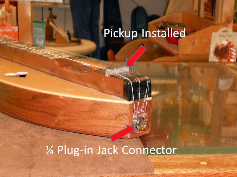 The Pickup Installation in a New Mountain Dulcimer Only features a musical instrument equipped with an installed pickup and a ¼ plug-in jack connector, highlighted with arrows.