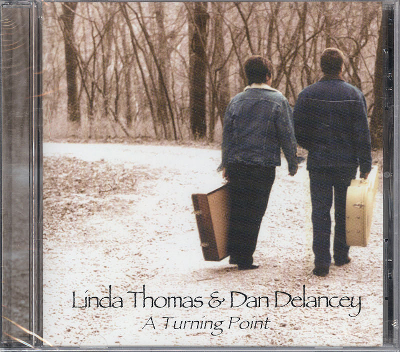 Two individuals walking down a wooded path, carrying suitcases, on the cover of A Turning Point - Linda Thomas and Dan DeLancey.