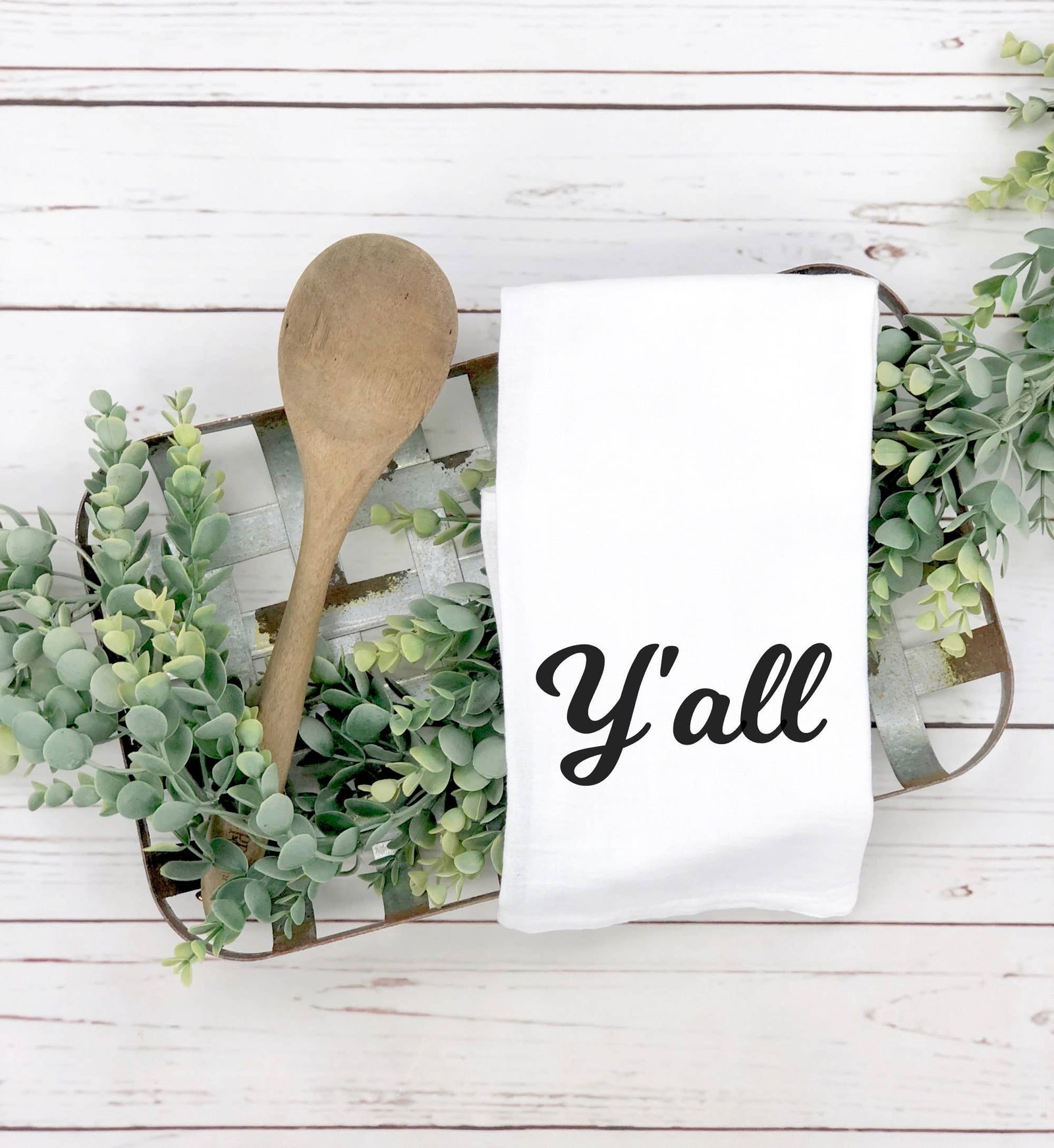A discontinued Y'all Tea Towel and a wooden spoon rest on a tray with green faux foliage atop a white wood surface—ideal as Southern kitchen decor or a thoughtful housewarming gift.