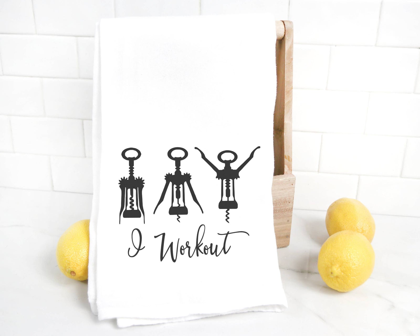 The "I Workout Tea Towel DISCONTINUED" is a white luxury cotton towel featuring black corkscrew illustrations and the phrase "I Workout"—a humorous tea towel, ideal for wine lovers. Three lemons are shown beside the towel.