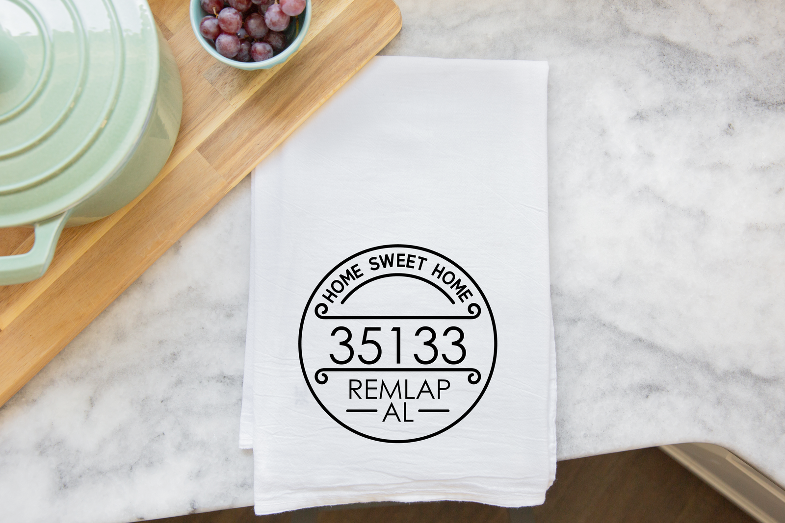 The discontinued Zip Code Tea Towel, printed with "HOME SWEET HOME 35133 REMLAP AL," rests on a marble counter beside a tray of grapes and a green dish—a personalized gift with your custom zip code.