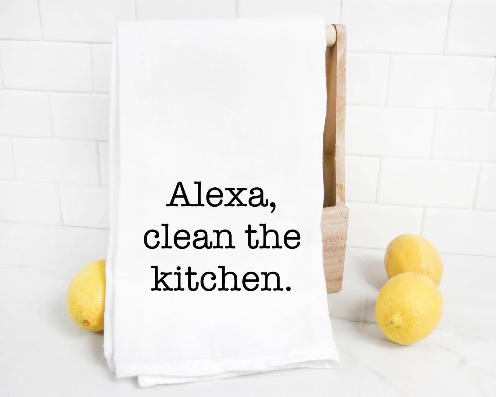 The discontinued “Alexa Clean the Kitchen Tea Towel” is shown on a white countertop beside three lemons, with a tiled backsplash in the background.
