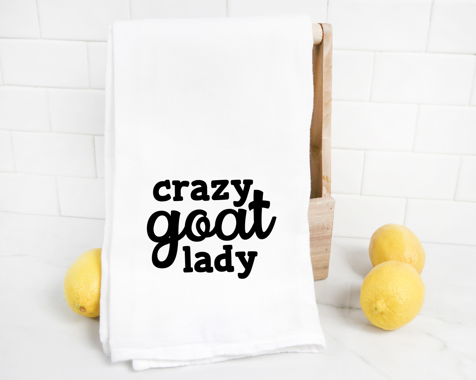The Crazy Goat Lady Tea Towel DISCONTINUED features "crazy goat lady" in black on white, shown on a wooden holder with lemons—an ideal quirky gift for goat lovers to brighten any kitchen counter.
