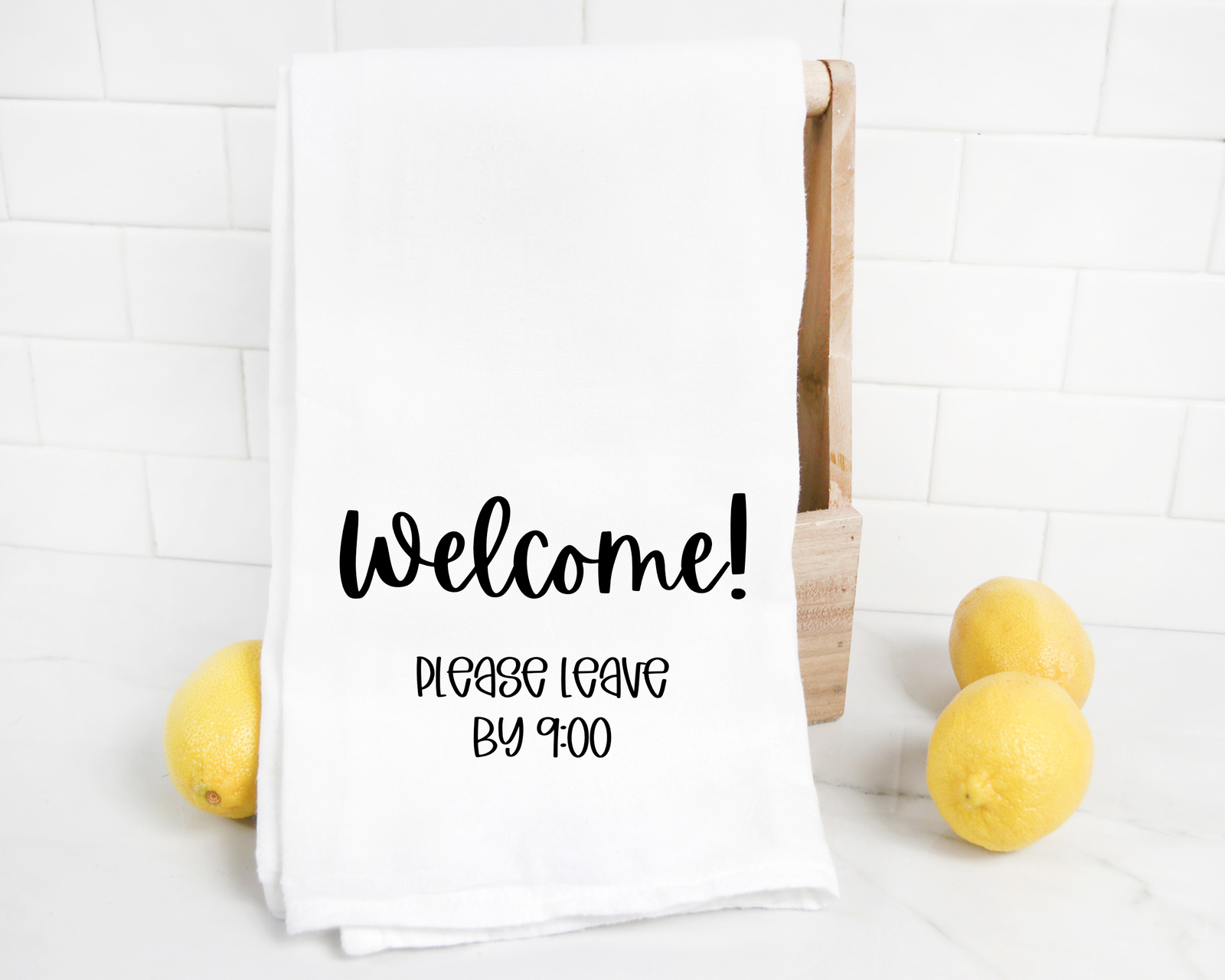 The discontinued "Welcome Please Leave By 9 Tea Towel" is displayed on a wooden holder with three lemons on a white countertop—a fitting gift for introverts who appreciate their alone time.